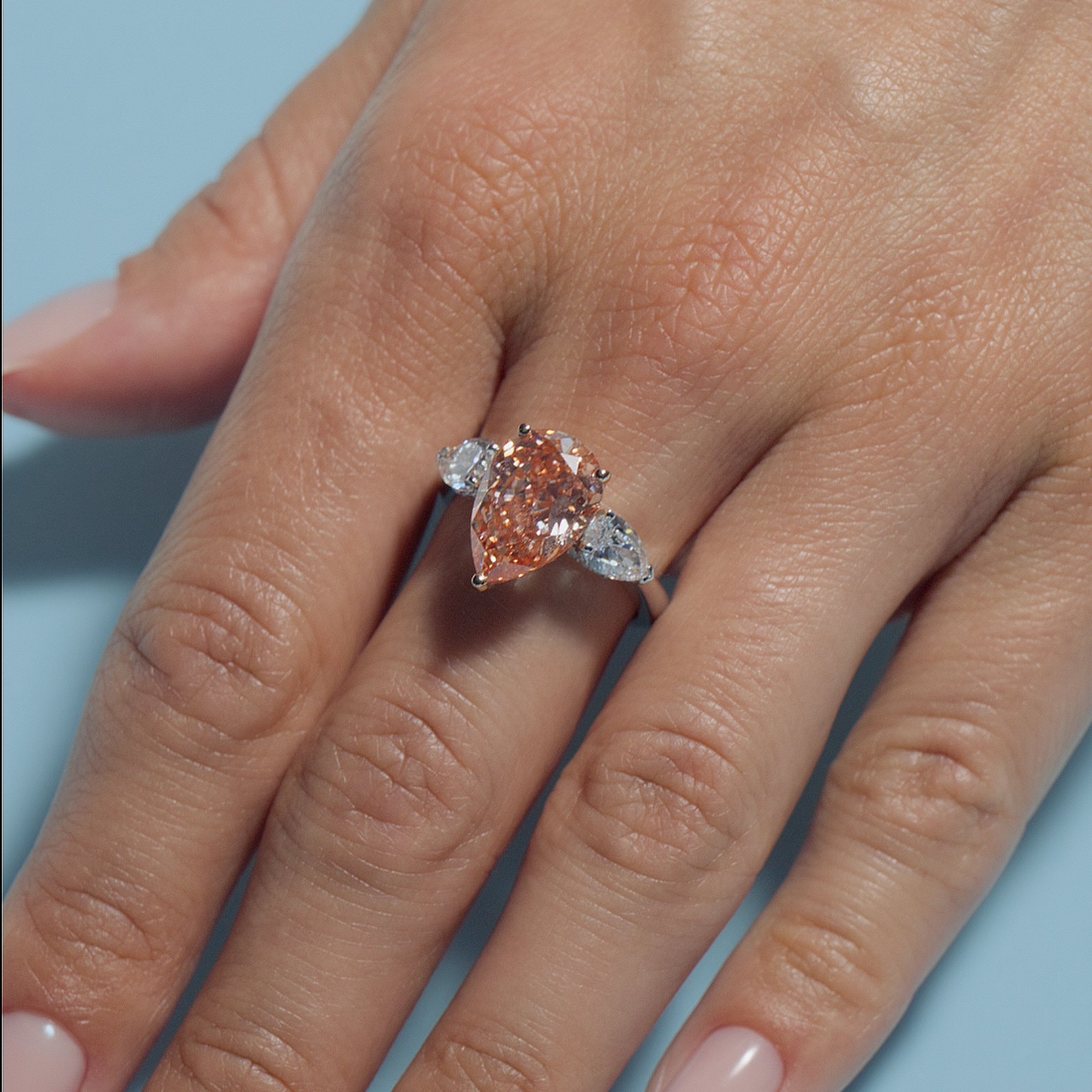Pear Fancy Vivid Orangy Pink Lab Diamond Pear Trilogy Ring on a hand against a light blue background