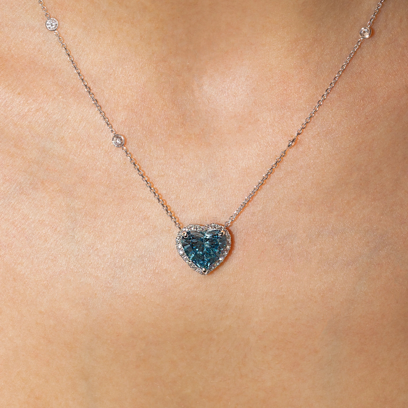 Heart-shaped blue lab-grown diamond halo pendant necklace in white gold with sautoir, worn on the neck.