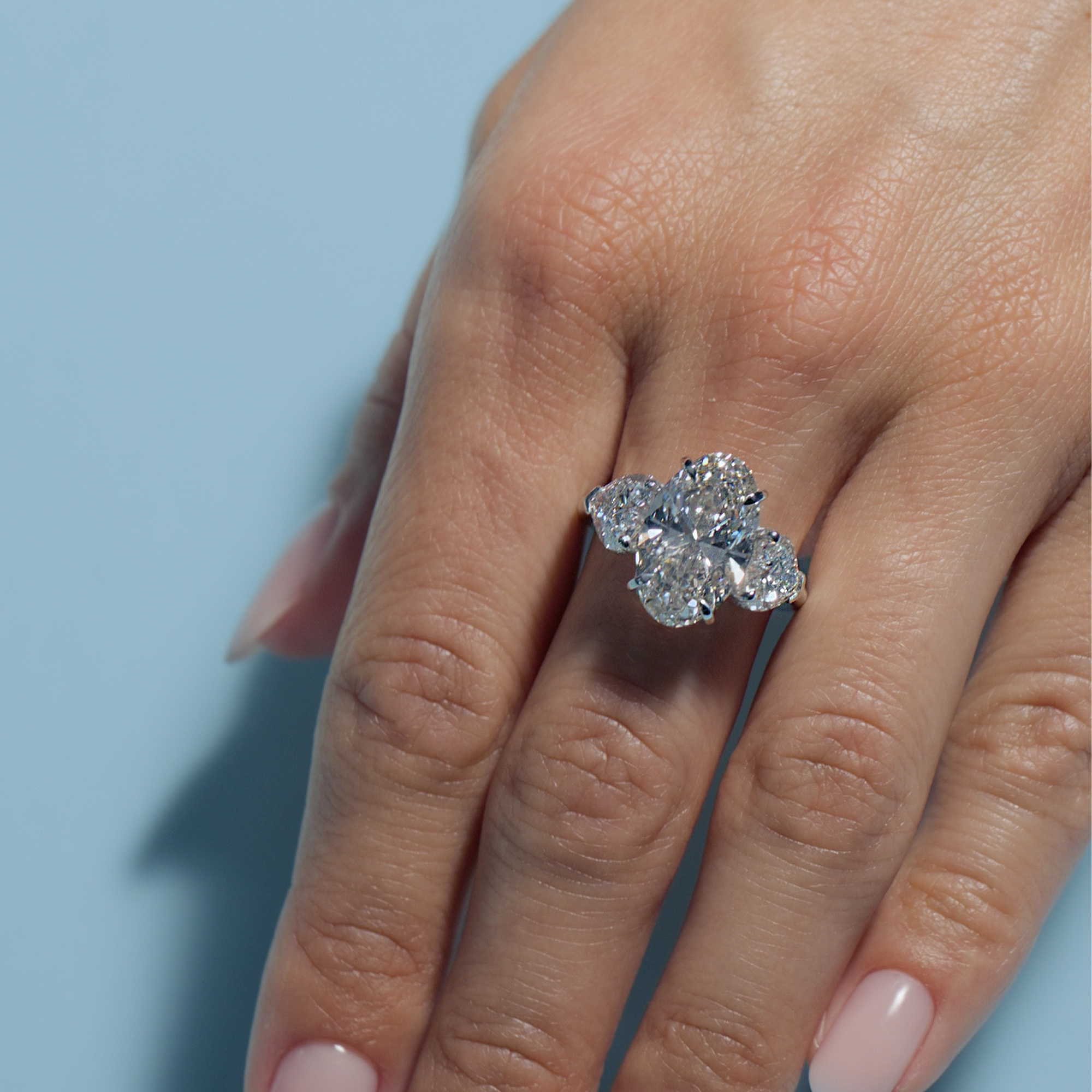 Close-up of a hand wearing a 5.04ct Oval Lab Diamond Heart Trilogy against a light blue background
