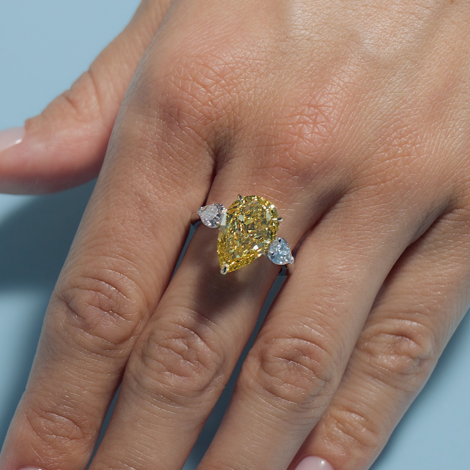 Close-up of a hand wearing a 5.6ct Fancy Vivid Yellow Pear Lab Diamond Classic Trilogy Ring on a light blue background