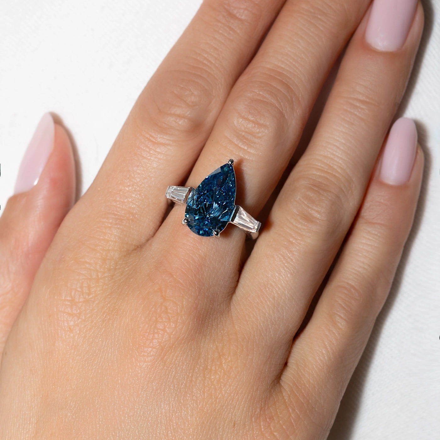 Pear-cut blue lab diamond ring with tapered baguette side diamonds worn on a hand against a white background