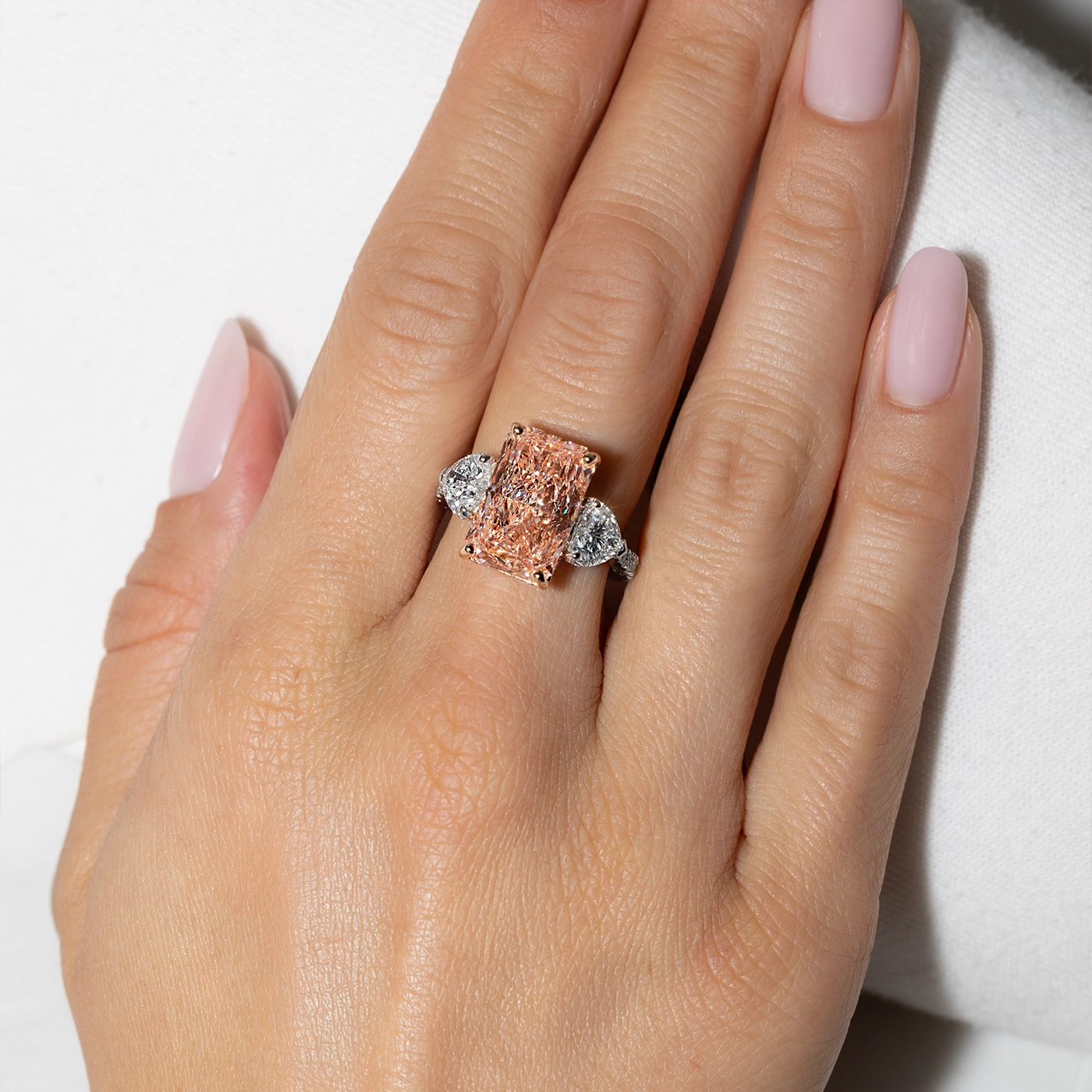 Radiant-cut pink diamond ring with heart-shaped side stones worn on a hand against a white background