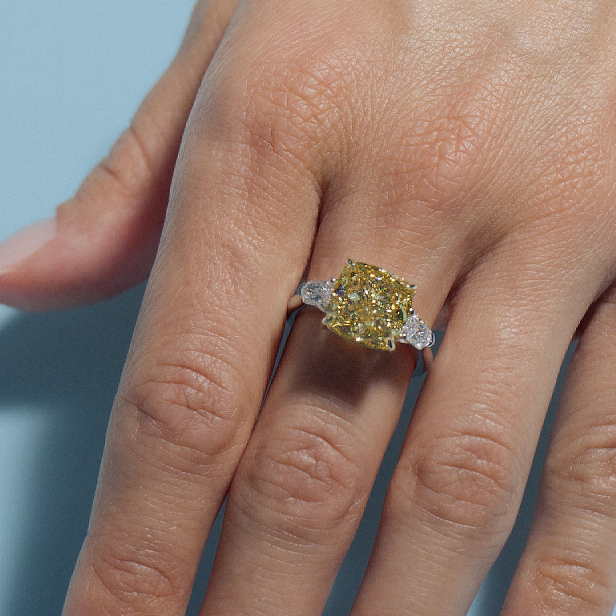 Close-up of a hand wearing a 6.2ct Fancy Vivid Square Cushion Lab Diamond Classic Trilogy Ring on a light blue background