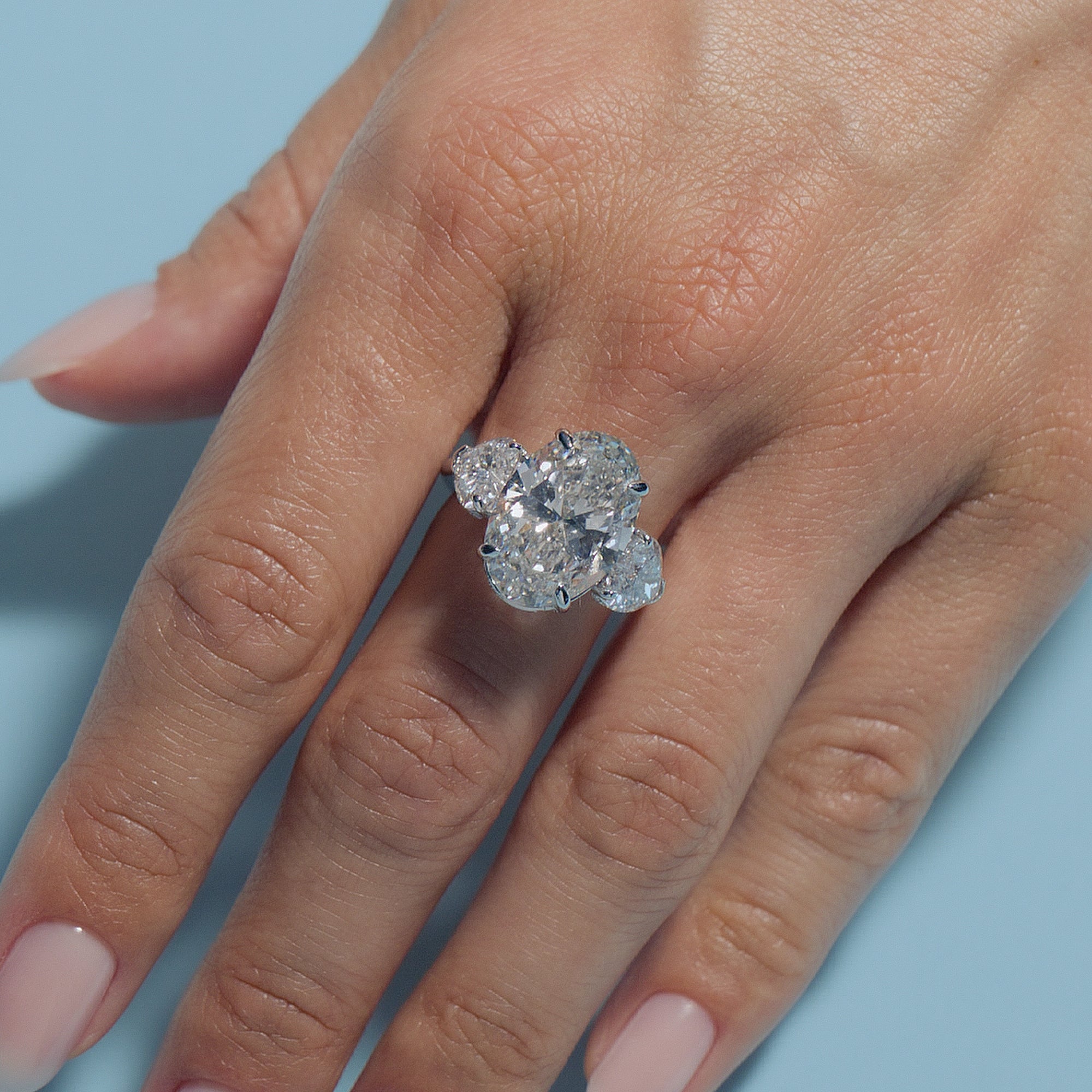 Close-up of a hand wearing a Oval Lab Diamond Heart Trilogy ring on a light blue background