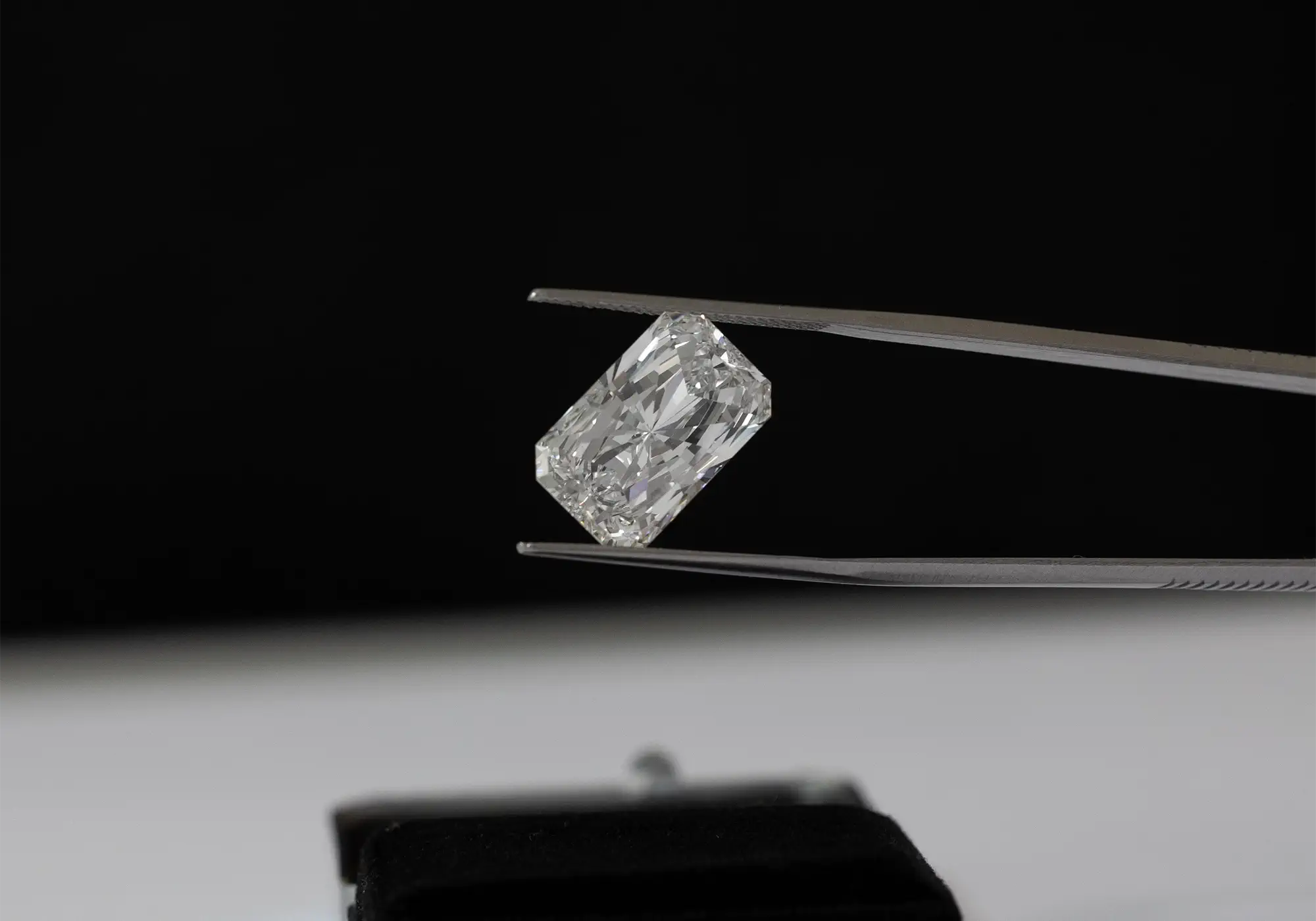 Radiant-cut diamond held by tweezers against a dark background on a jeweler’s workbench