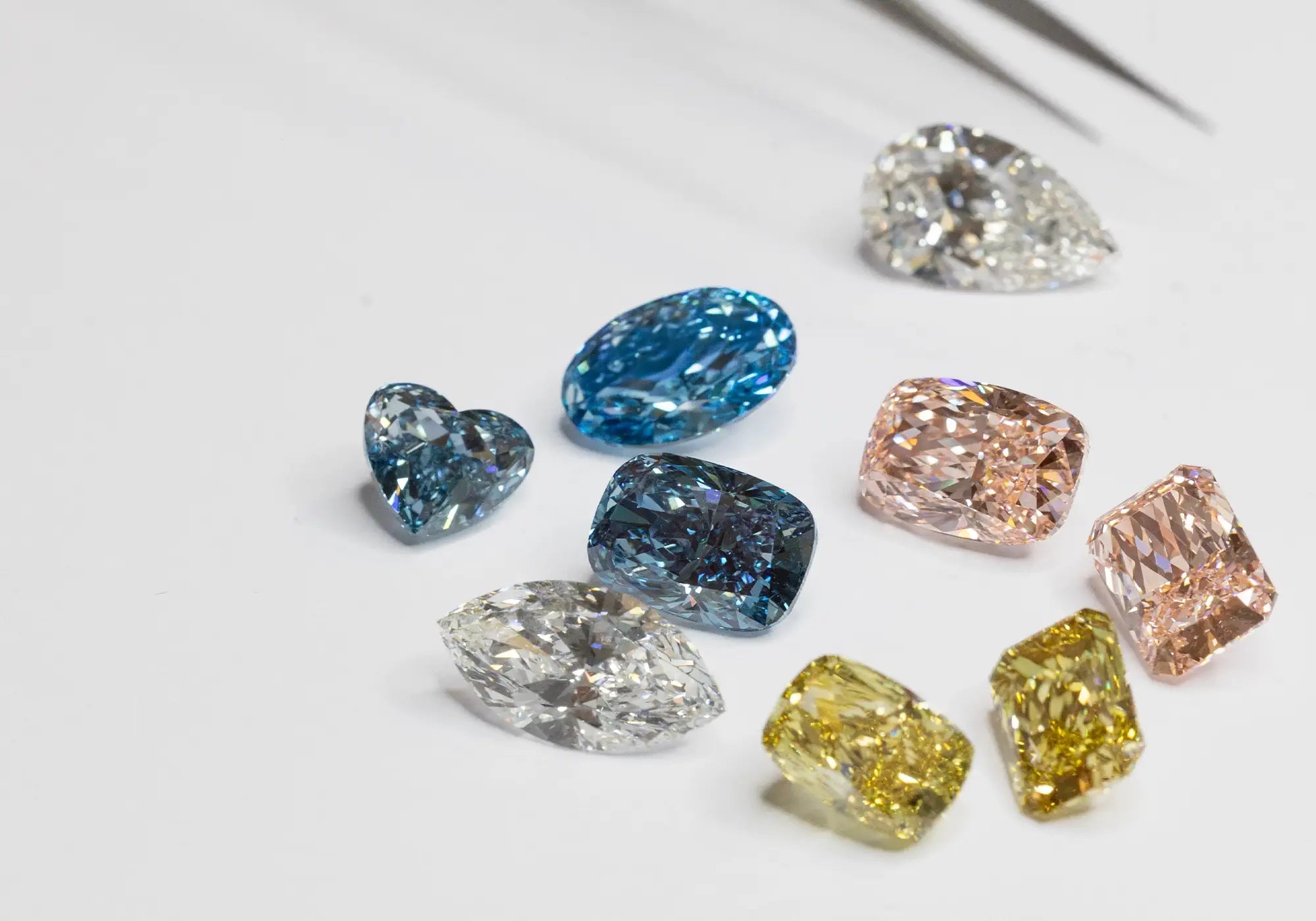 A mix of colorful, cut lab-grown diamonds are spread out on a white surface, catching the light. Tweezer is place at the back