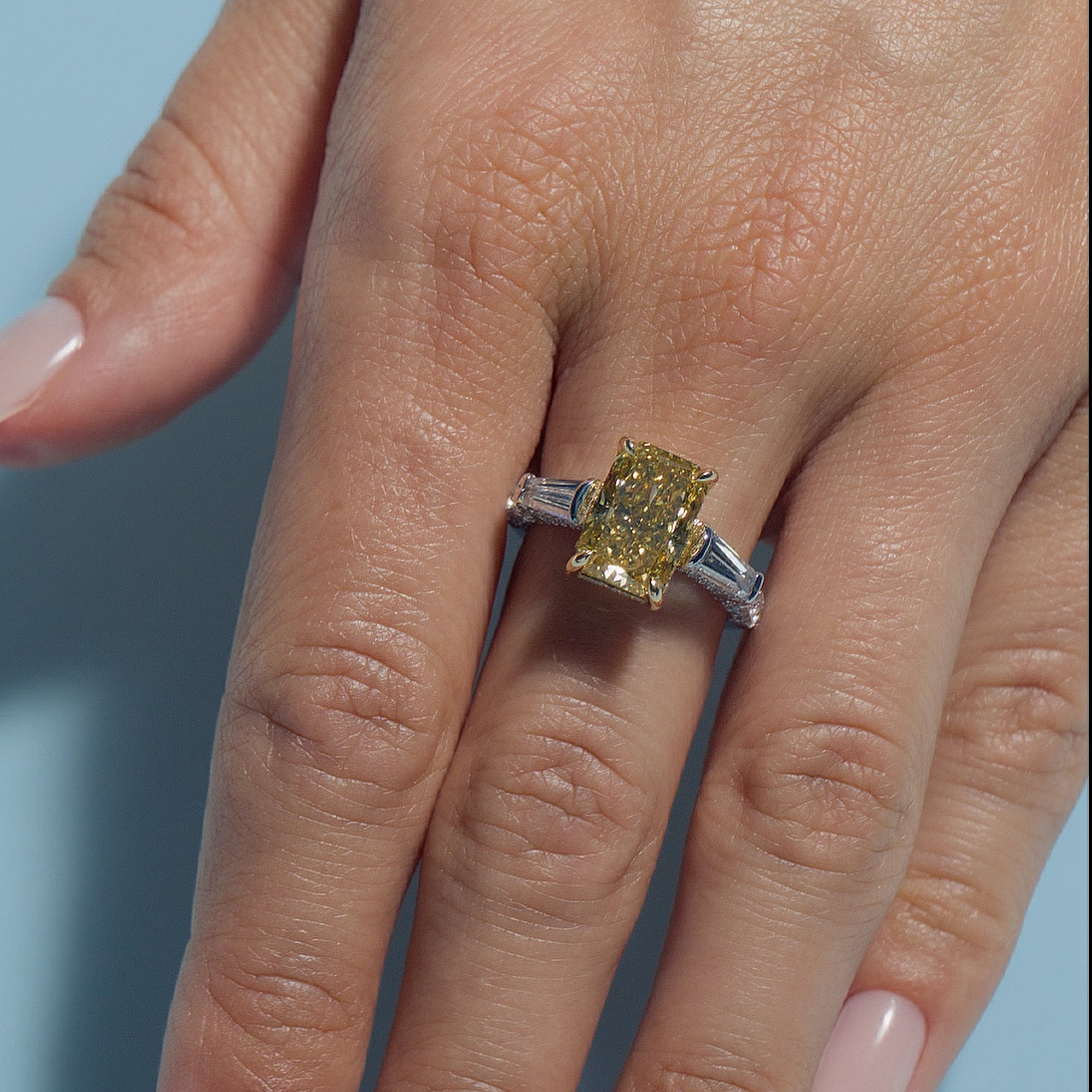 Close-up of a hand wearing a Radiant Fancy Vivid Yellow Lab Diamond Tapers Trilogy ring with a light blue background