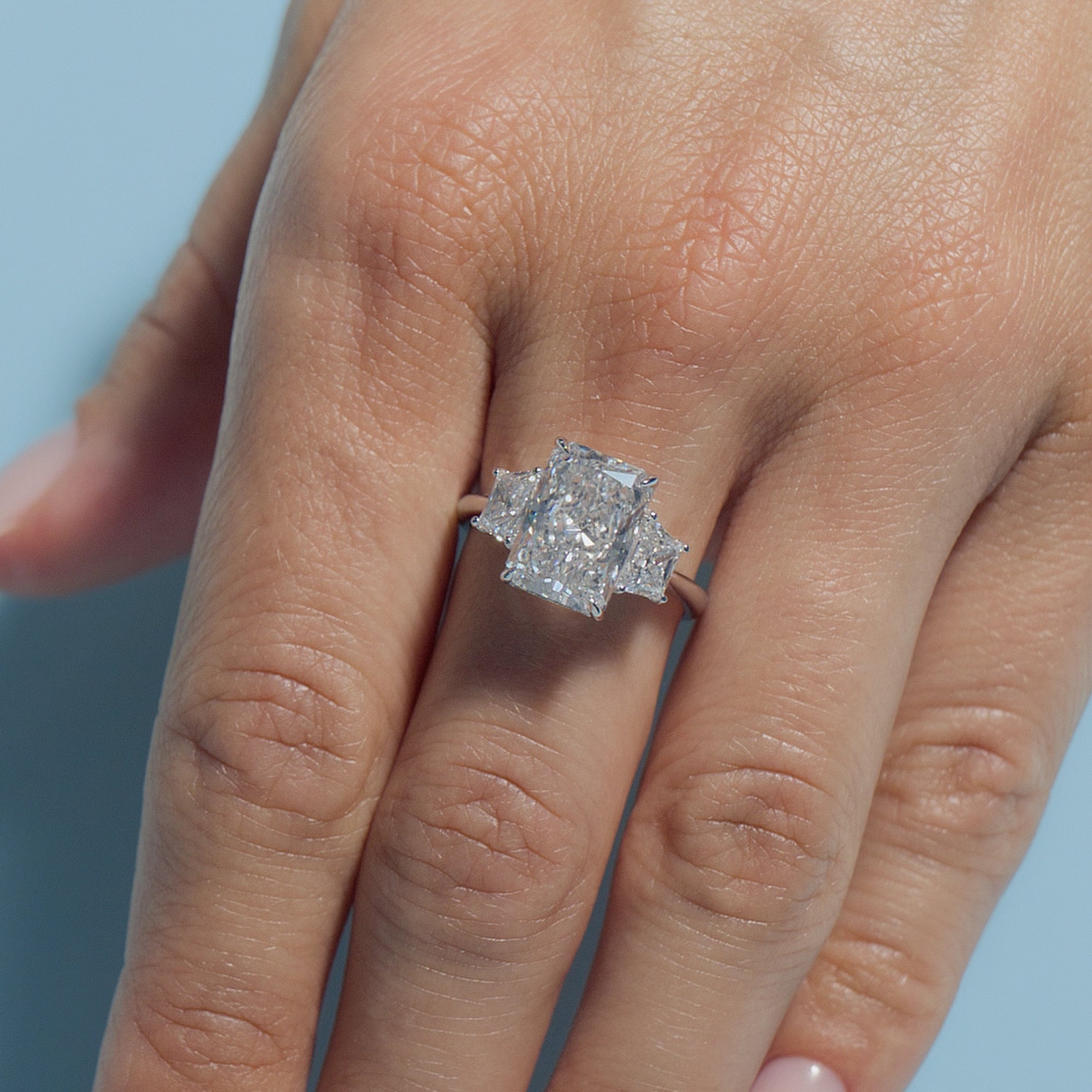 4.06ct Radiant Lab Diamond Classic Trilogy ring on a finger against a light blue background