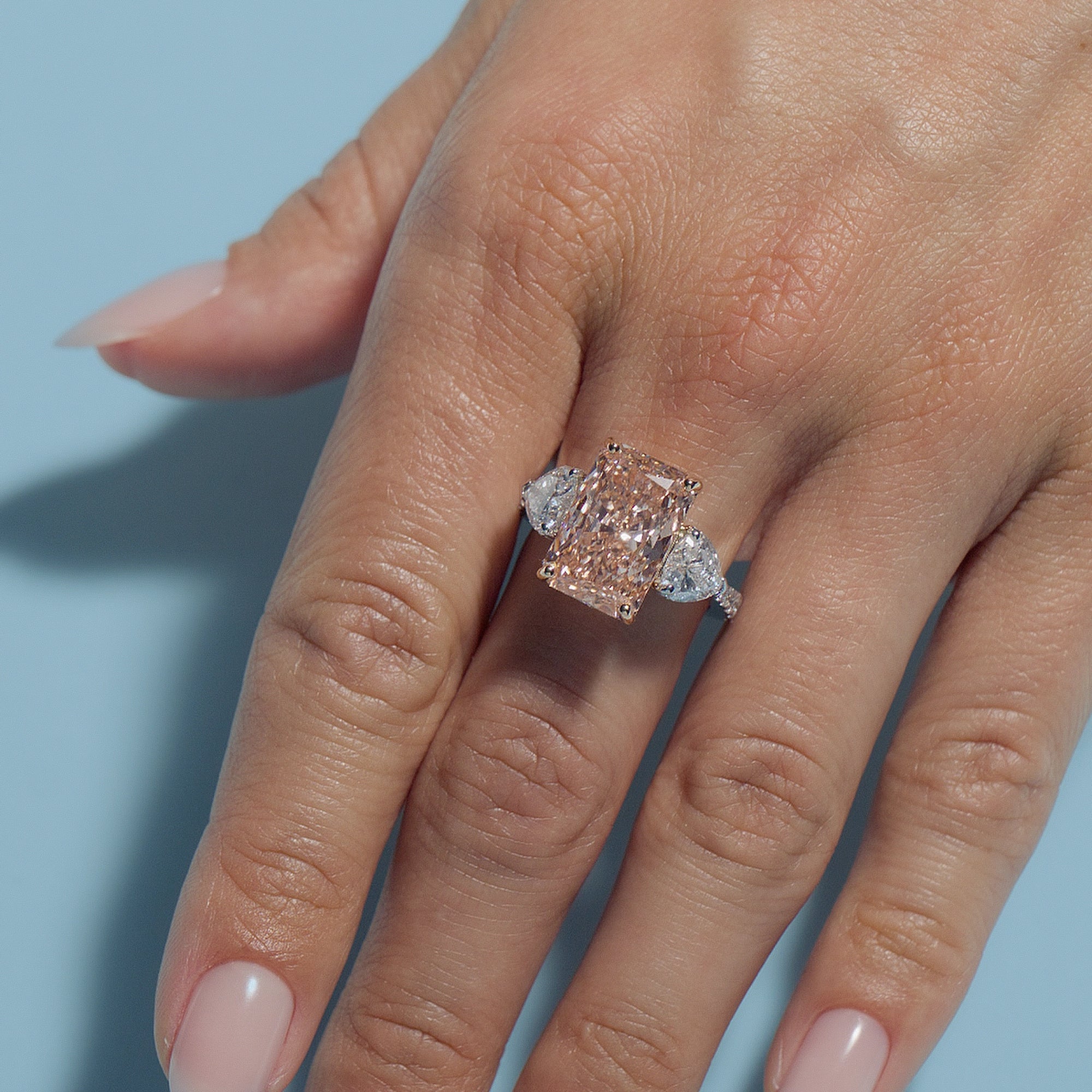 Hand wearing a Radiant Fancy Vivid Pink Lab Diamond Heart Trilogy ring with a light blue background