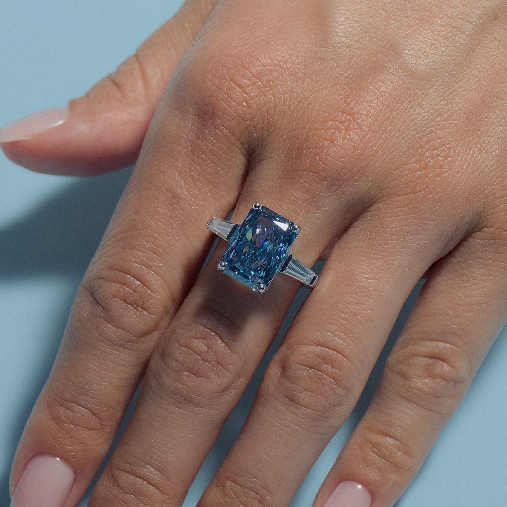Radiant Fancy Vivid Blue Lab Diamond Tapers Trilogy ring on a hand against a light blue background