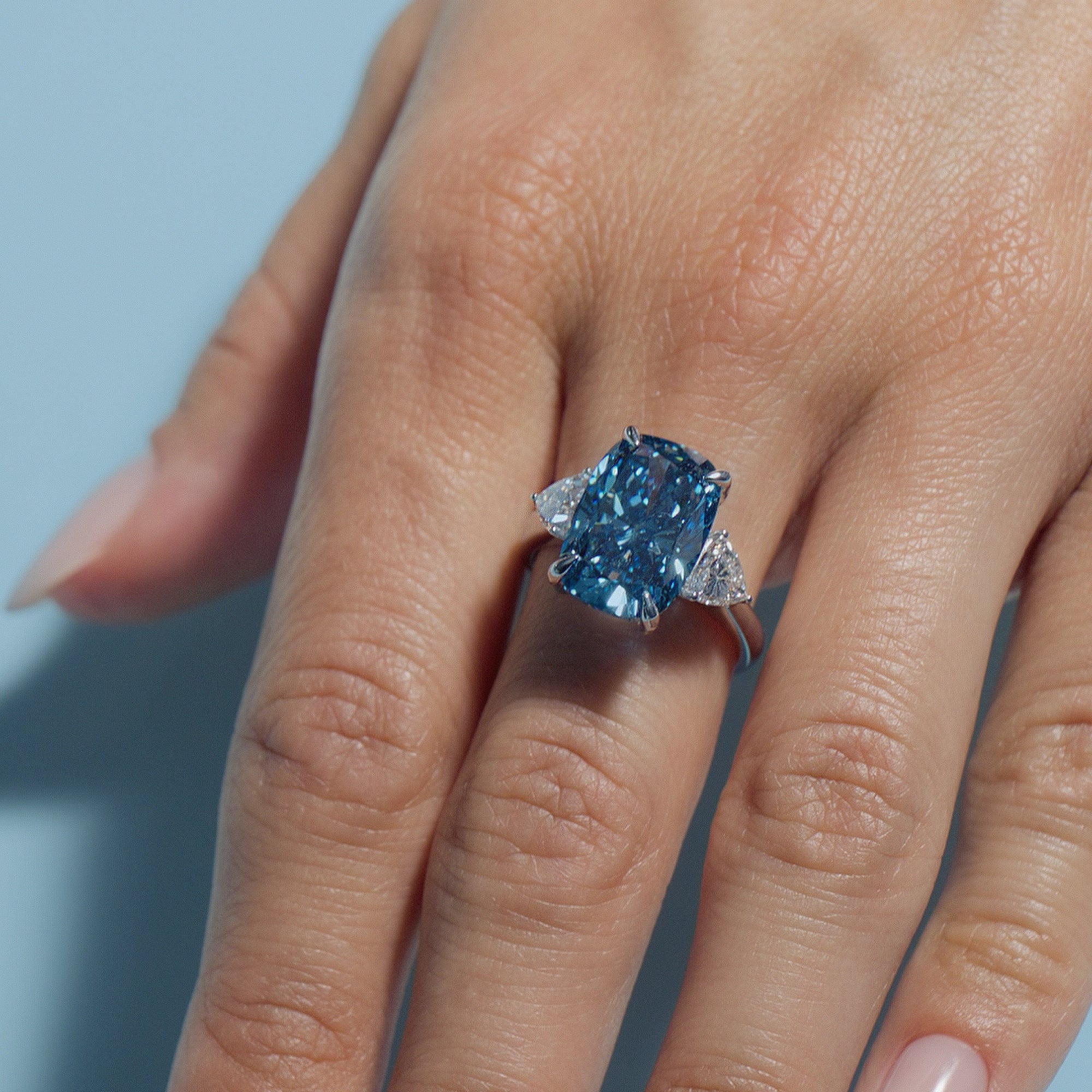 7.04ct Fancy Vivid Blue Cushion Lab Diamond Classic Trilogy ring worn on a hand against a light blue background