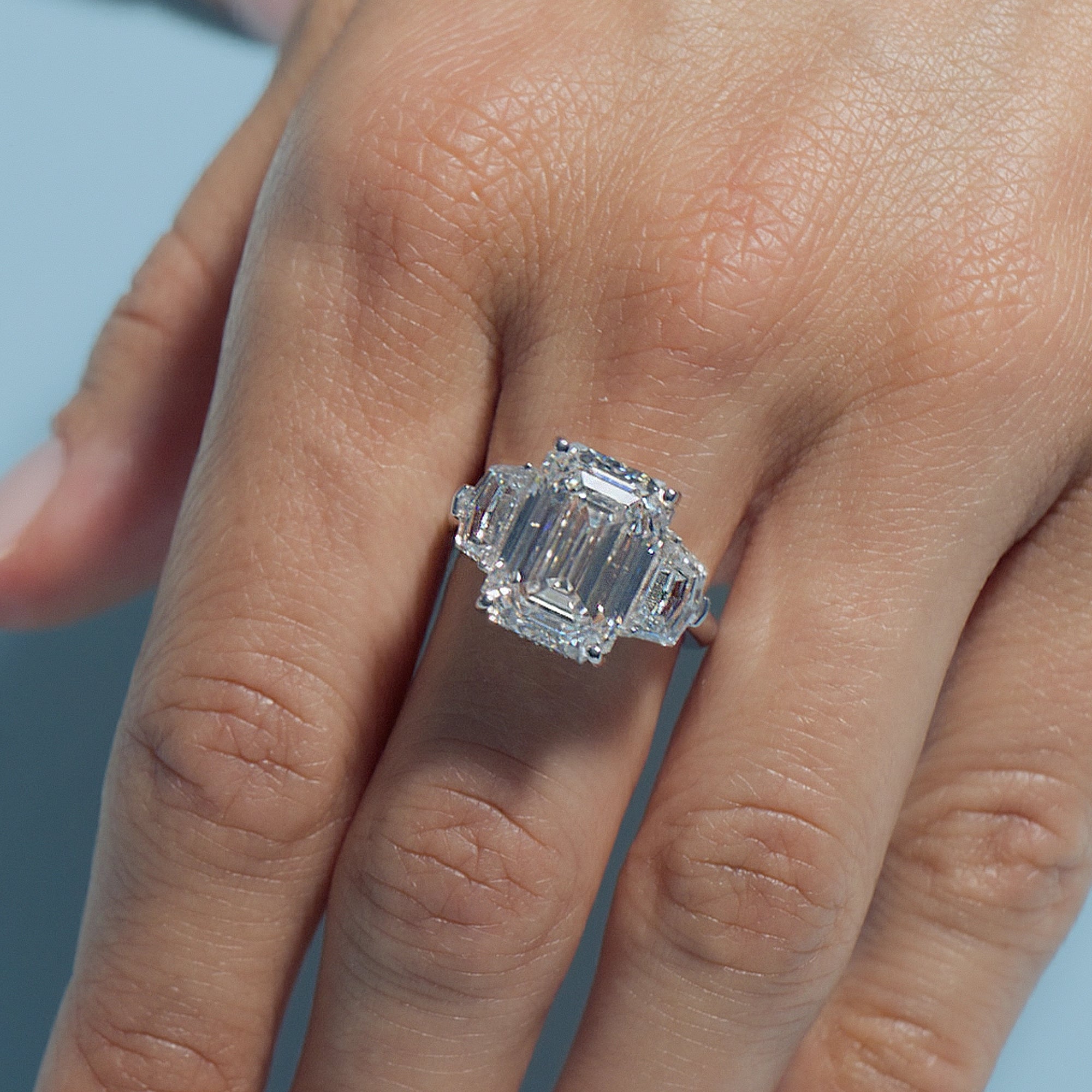8.01ct Emerald Lab Diamond Intricate Trilogy ring on a finger against a light blue background