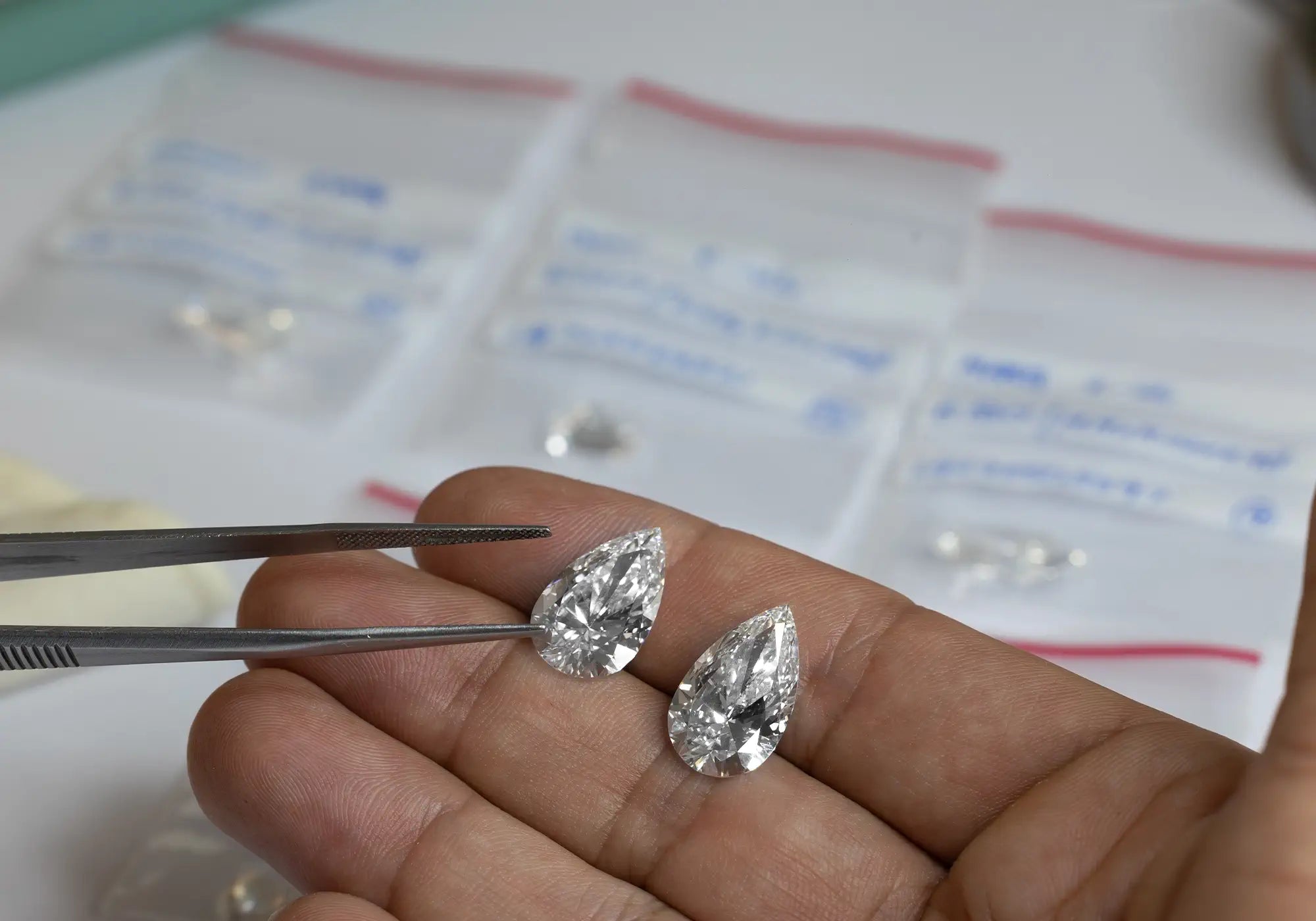 Two pear-shaped diamonds are held with tweezers over a hand. Small labeled bags in the background with lab-grown diamonds inside it also. 