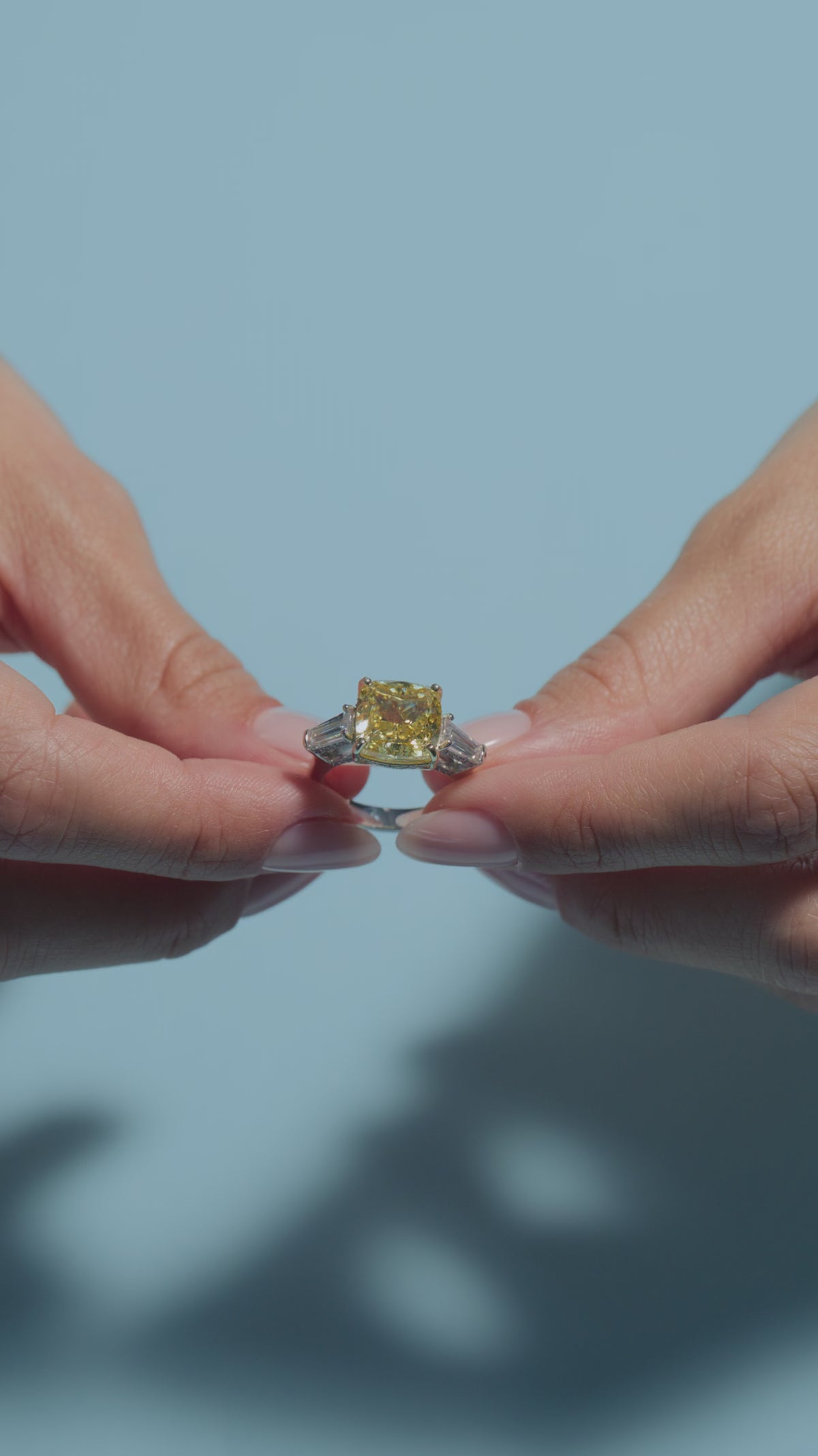 A video clip presenting a 6.7ct Fancy Vivid Yellow Square Cushion Lab Diamond Intricate Trilogy Ring held and worn by the model on a light blue background