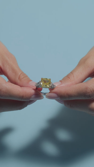 A video clip presenting a 6.7ct Fancy Vivid Yellow Square Cushion Lab Diamond Intricate Trilogy Ring held and worn by the model on a light blue background