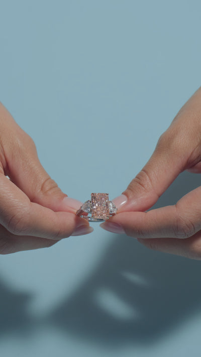 A video clip  of a 6.08ct Radiant Fancy Vivid Pink Lab Diamond Heart Trilogy Ring shown off by the model of BAL Diamonds on a light blue background