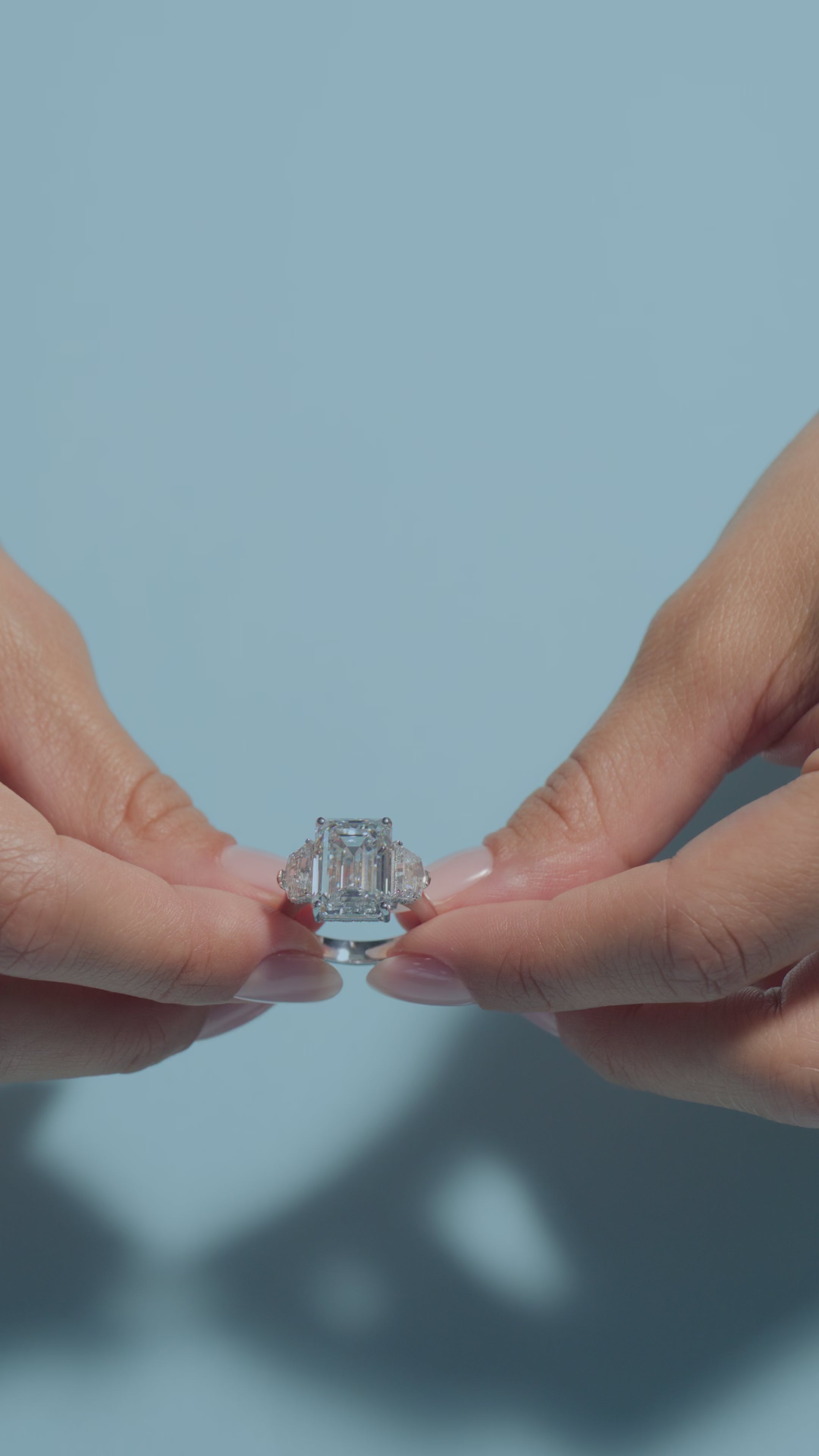 A vide presenting a 8.06ct Emerald Lab Diamond Intricate Trilogy Ring held and worn by the model on a light blue background