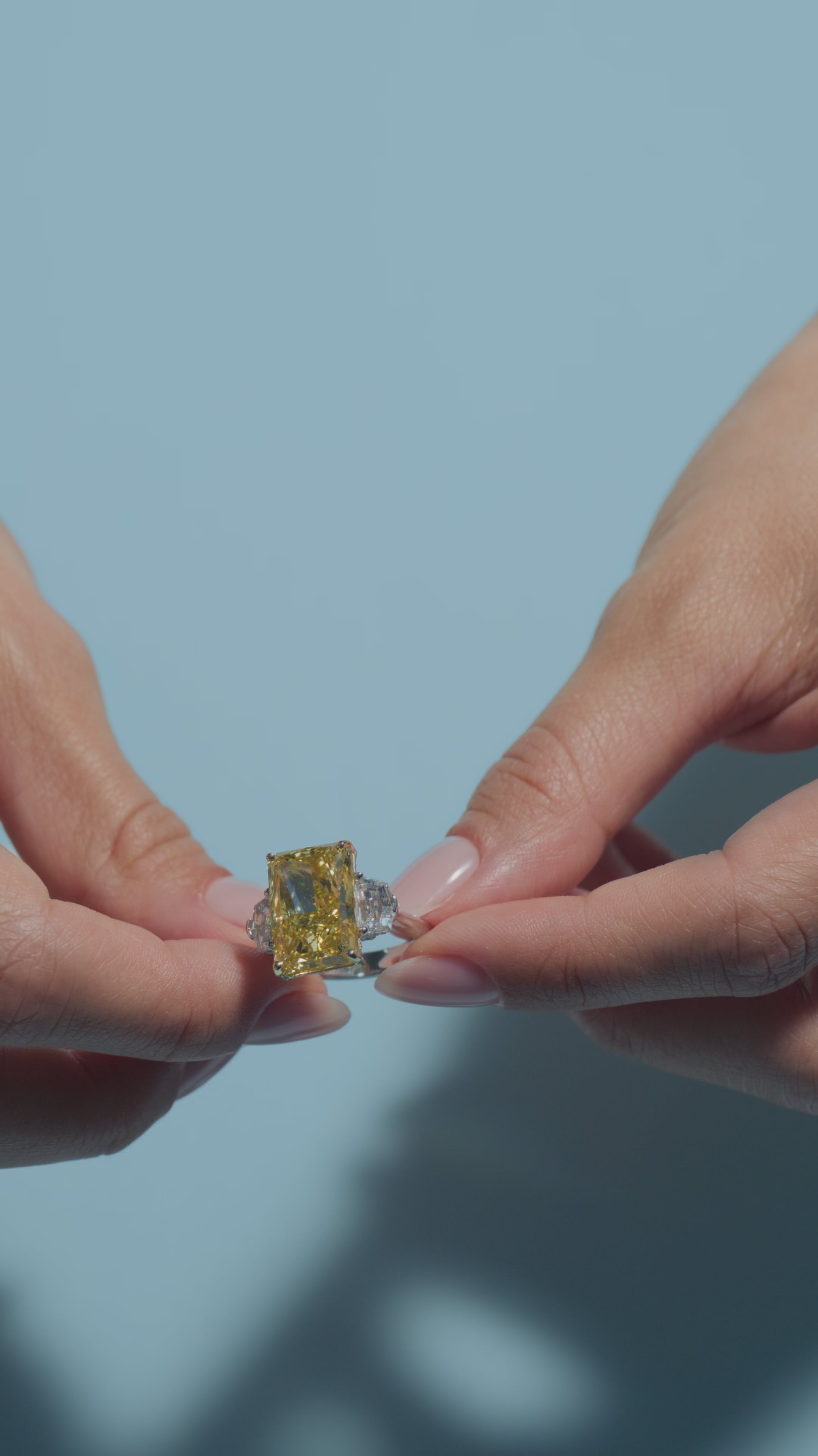 A video presentation of a 11.2ct Fancy Vivid Yellow Radiant Lab Diamond Intricate Trilogy Ring held and worn by the model of BAL Diamonds on a light blue background