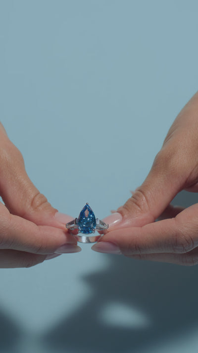 A short video featuring a 5ct Pear Fancy Vivid Blue Lab Diamond Tapers Trilogy Ring held by the  model on a light blue background 